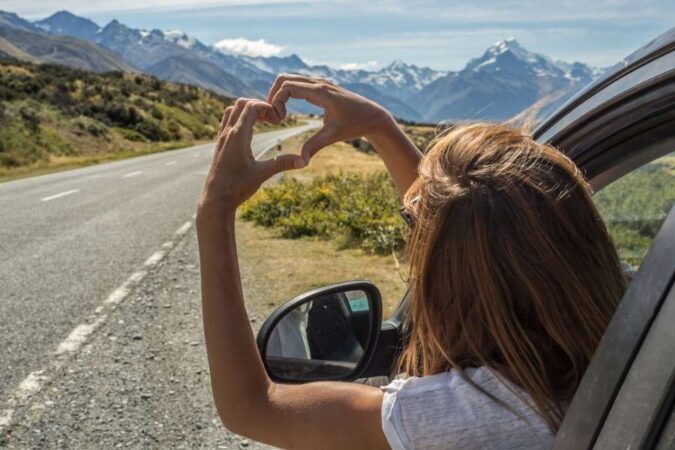 Une femme forme un cœur avec ses mains depuis la fenêtre d'une voiture face à une route de montagne, l'essence du slow trip.