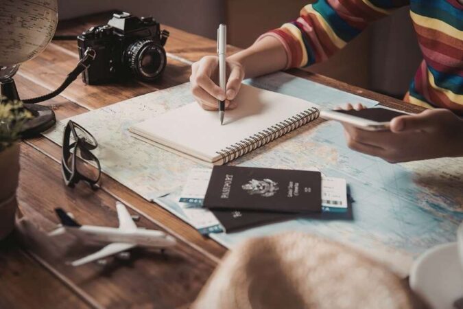 Une personne prend des notes détaillées dans un carnet ouvert sur une carte du monde pour organiser son circuit tout compris, entourée d'un passeport et d'un appareil photo.