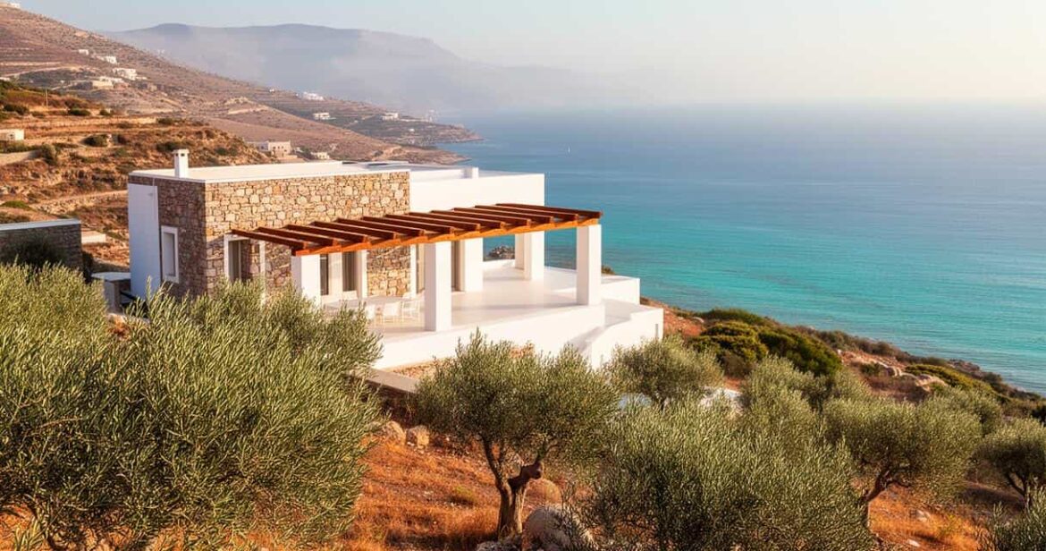 Vue d'une villa moderne en pierre et stuc blanc avec pergola, perchée sur une colline couverte d'oliviers, idéale pour des locations de villas en Crète pour vos vacances, surplombant la mer turquoise et des montagnes au loin.